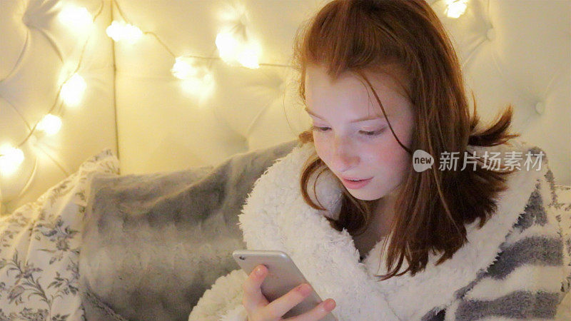 14 / 15岁的红发少女坐在卧室里，靠在一堆靠垫和枕头上，穿着毛茸茸的灰白条纹睡袍，看着手机，被彩灯照亮